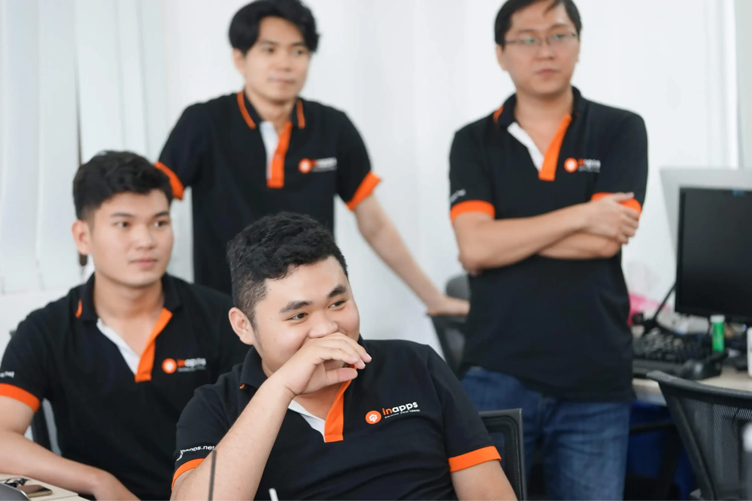 Four InApps engineers in branded polo shirts in the Ho Chi Minh City office, two seated in the foreground and two standing behind, sharing a laugh during a discussion